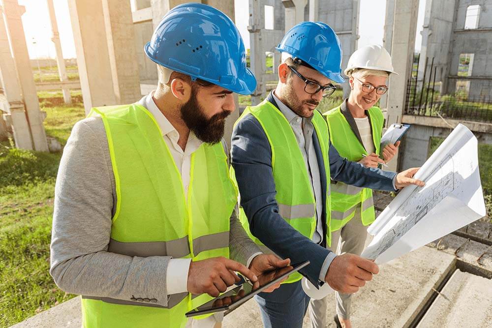 Project managers, two men and one woman, looking at building plans, wearing high visibility vests on construction site